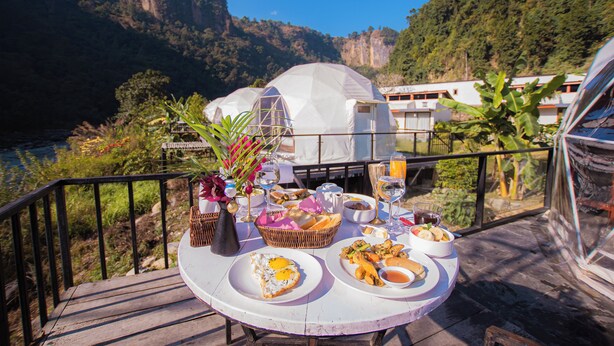 A dining table with breakfast items, a vase with flowers, and a view of a mountain and river. - Harness Zipline And Adventure Resort (Godaam)