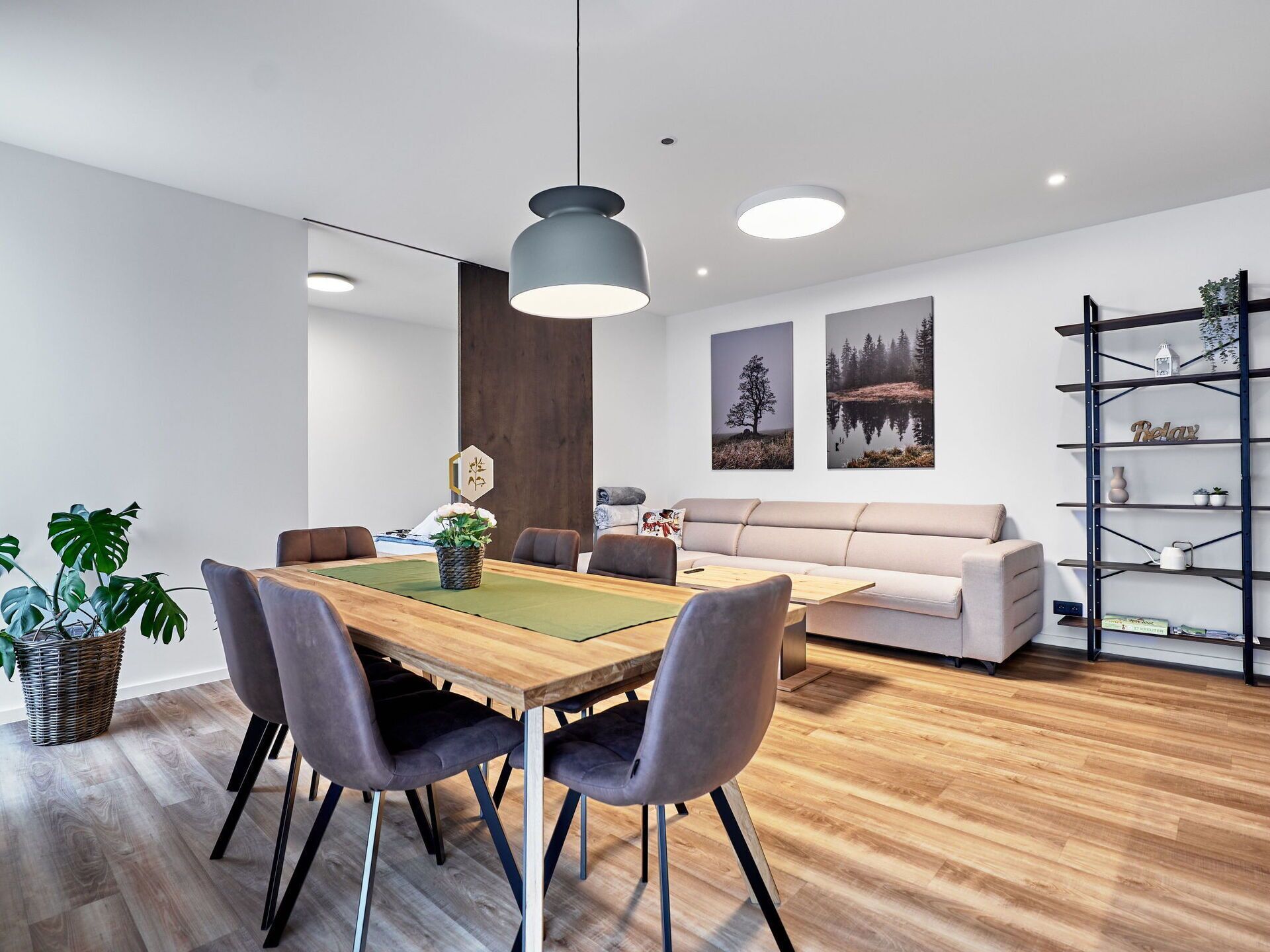 A modern living space with a wooden dining table, grey upholstered chairs, a beige sofa, and a black shelving unit with decorative items.