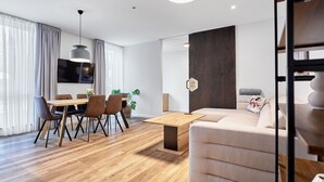 Een moderne woonkamer met houten vloer, een beige bank, een eettafel met stoelen en een aan de muur bevestigde televisie.