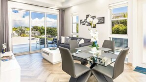 A modern living room with a glass dining table, grey chairs, a sofa, and a ceiling fan.
