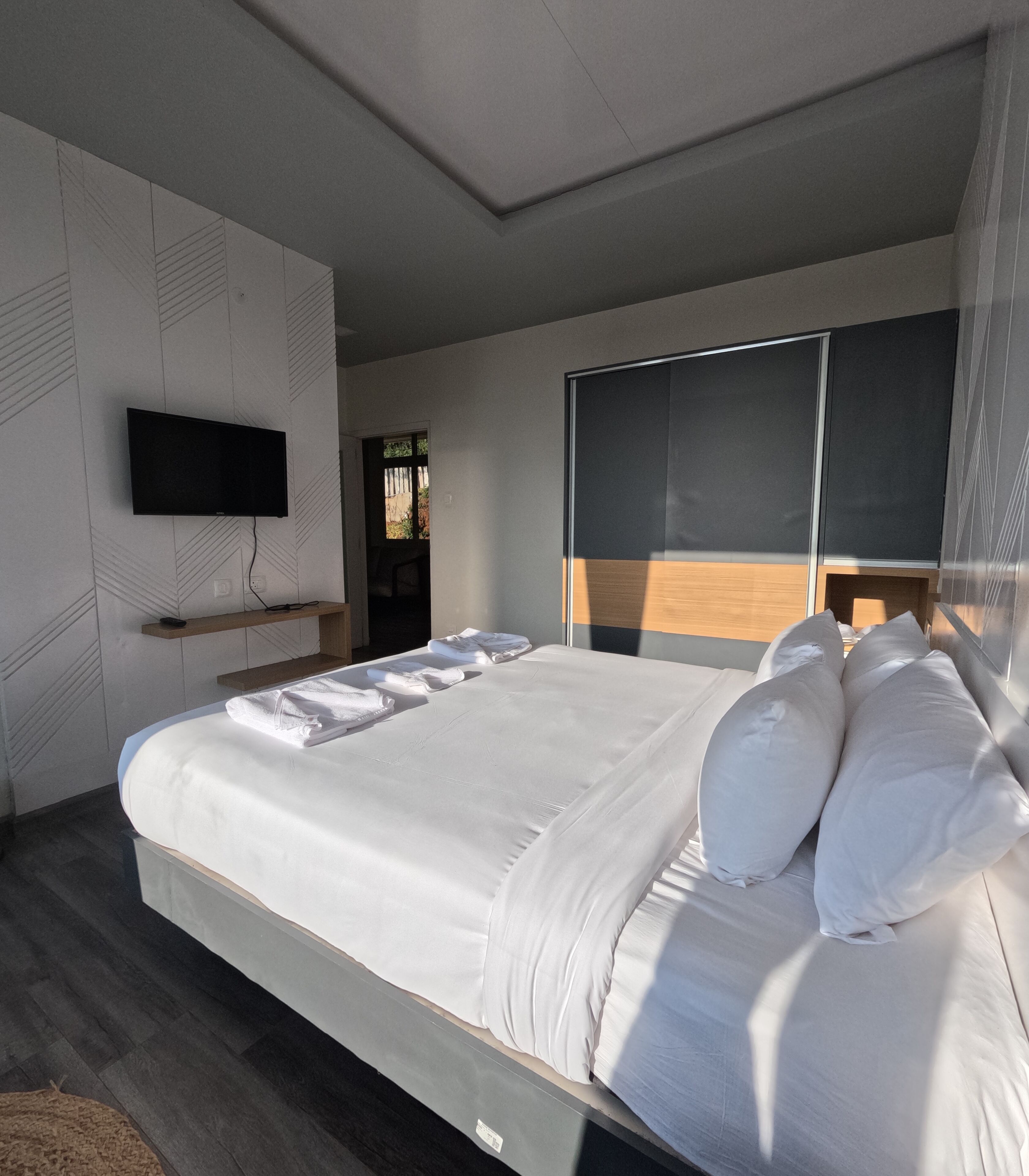A modern bedroom with a large bed, a flat-screen TV mounted on the wall, and a window with blinds.