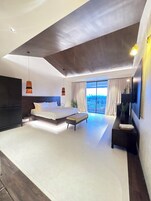 A modern bedroom with a large bed, a bench, a TV, and a view of the outdoors.