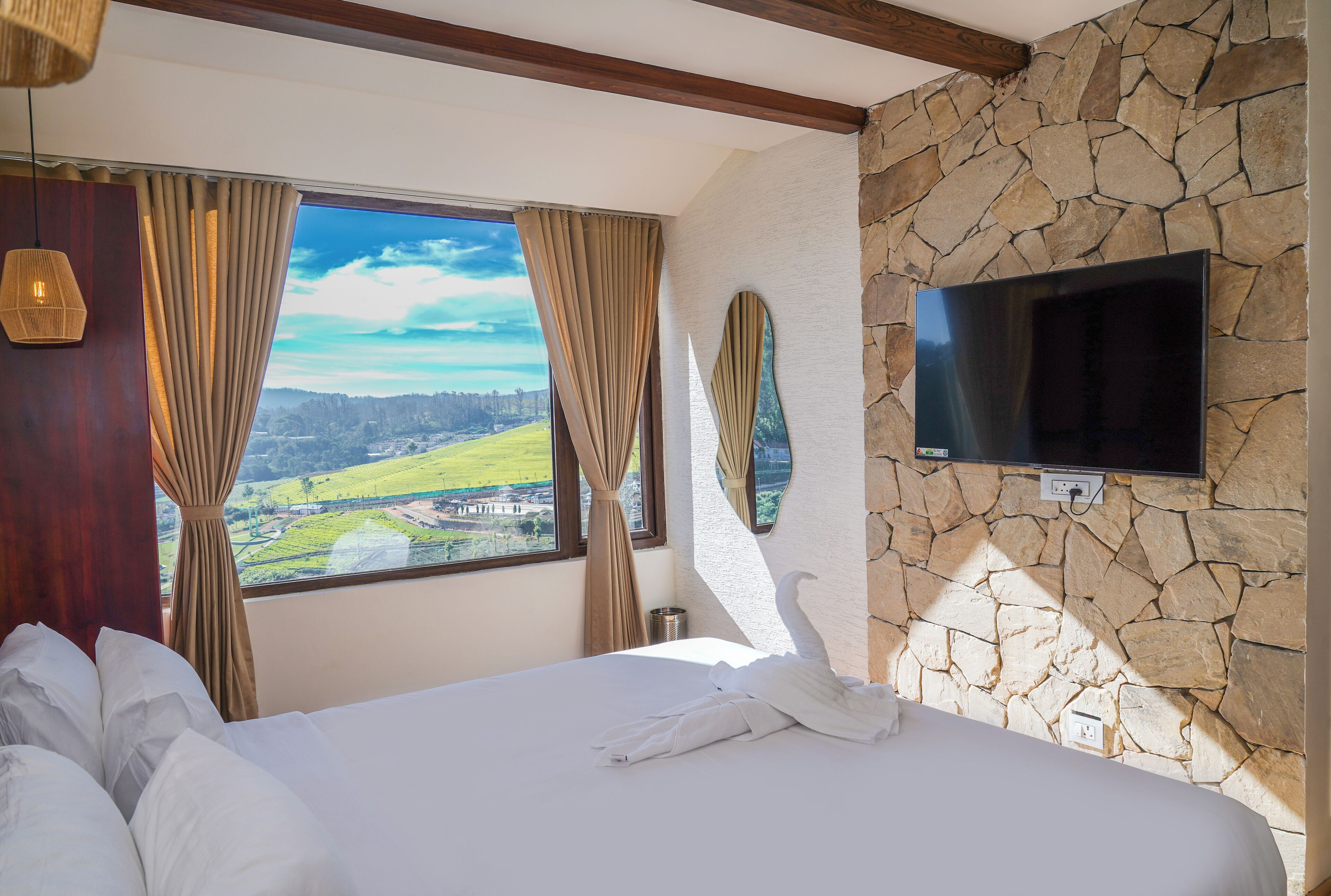 A hotel room with a large bed, a stone wall, a TV mounted on the wall, and a window with curtains.