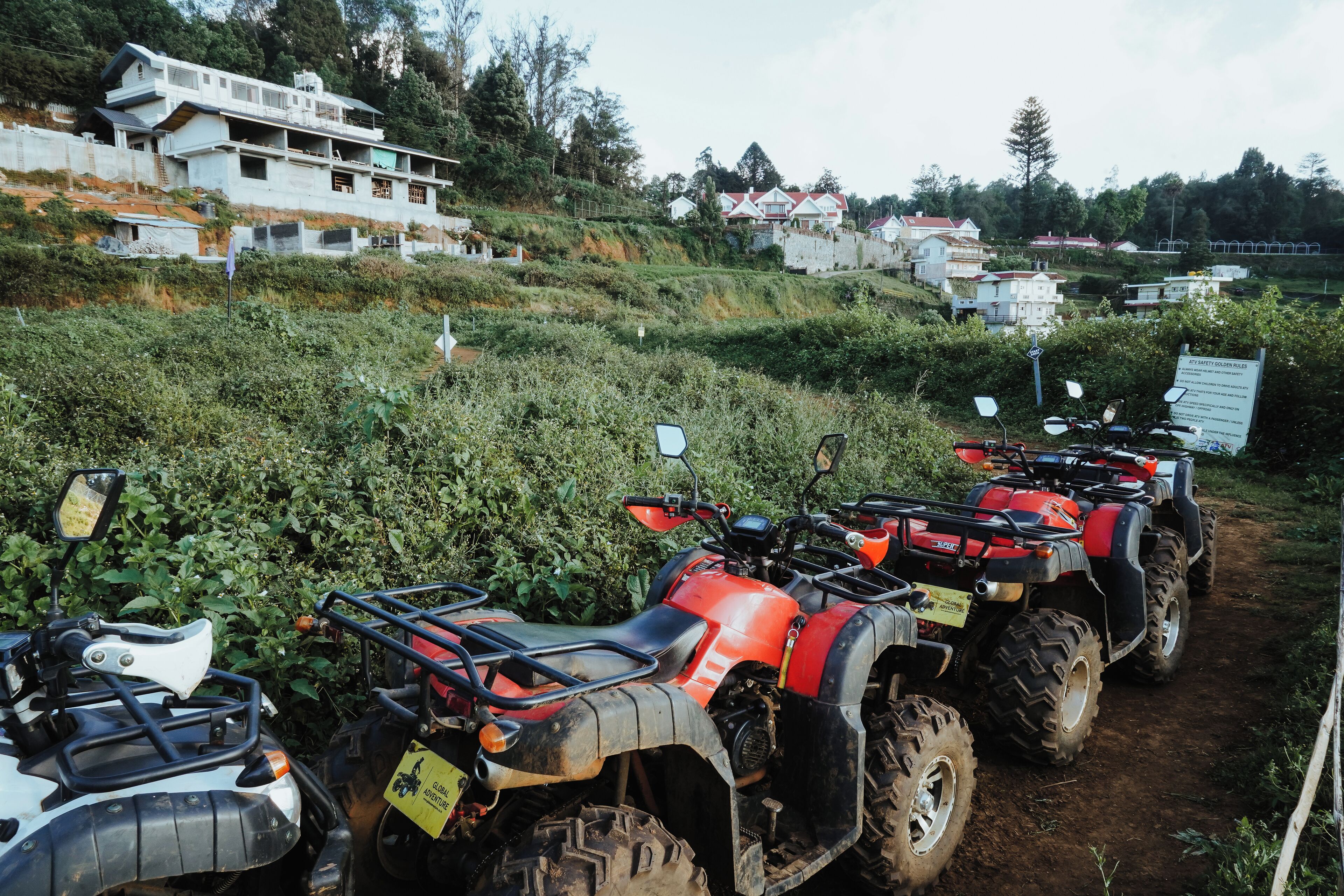 A row of ATVs parked on a dirt path with houses in the background.