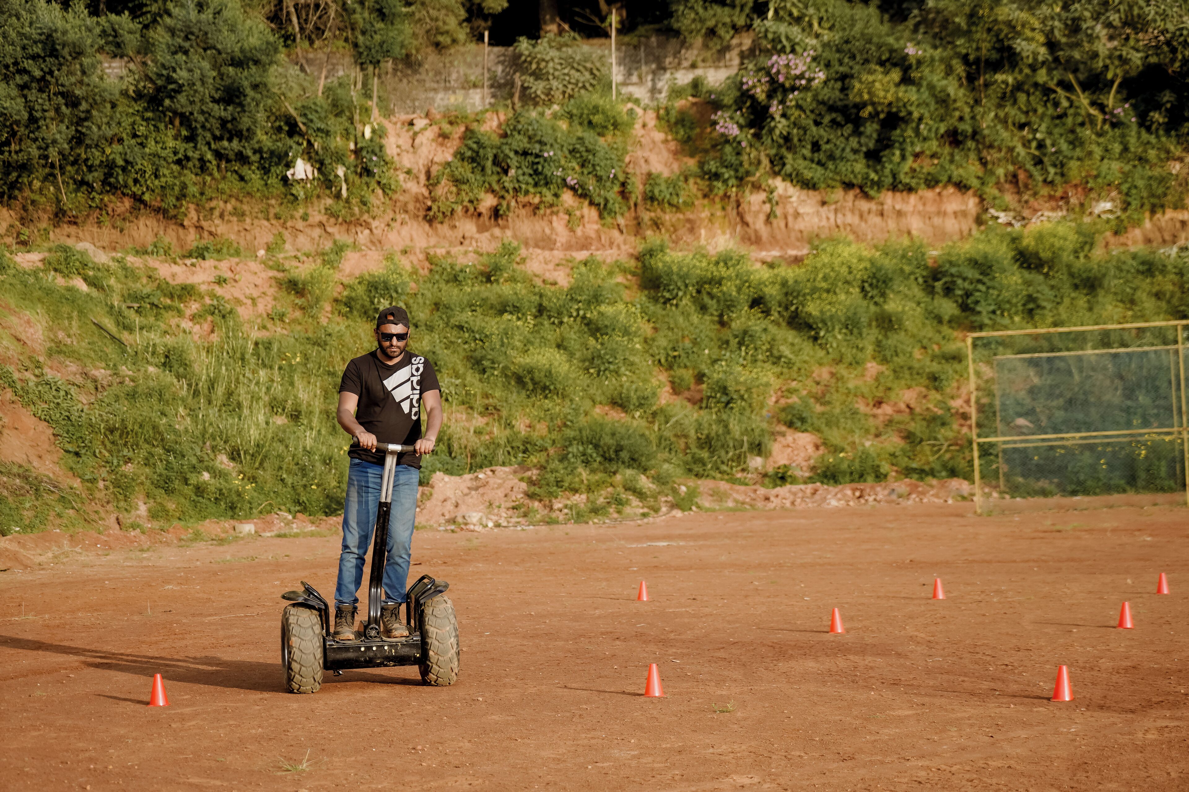 Une personne conduit un Segway sur un chemin de terre, délimité par des cônes orange, et est entourée de verdure et d’une clôture.