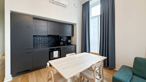 Une cuisine moderne avec des meubles de couleur sombre, un plan de travail blanc et un four encastré. On y trouve une table à manger avec quatre chaises et un canapé vert dans l’espace salon.