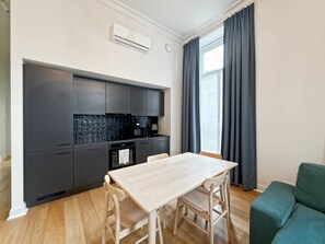 Eine moderne Küche mit dunklen Schränken, einer weißen Arbeitsplatte und einem eingebauten Ofen. Im Wohnbereich befindet sich ein Esstisch mit vier Stühlen und ein grünes Sofa.