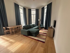 Ein modernes Wohnzimmer mit einem Ecksofa, einem Holzesstisch mit Stühlen und einem an der Wand montierten Fernseher.