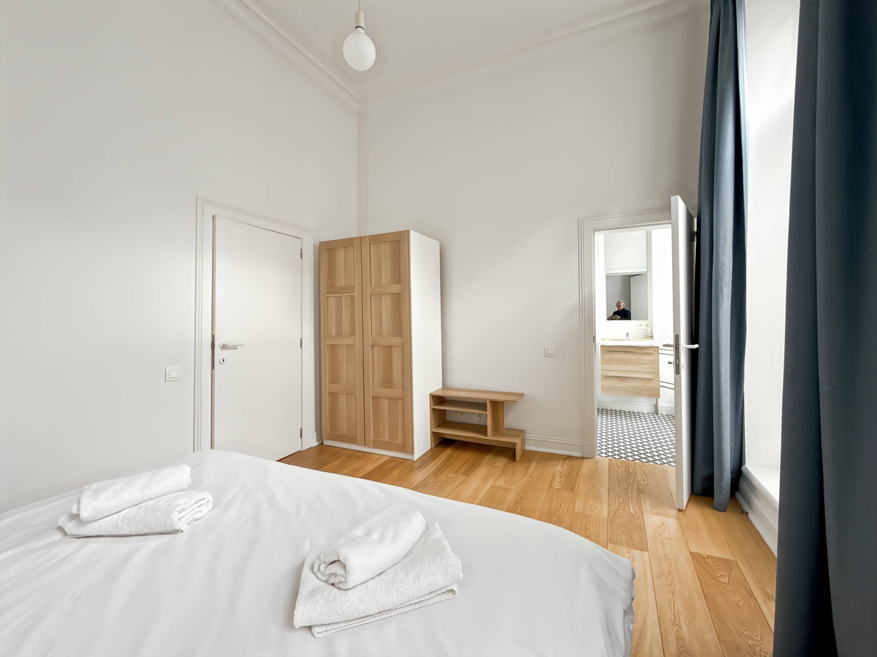 A modern bedroom with a large bed, a wardrobe, a small table, and a bathroom visible through an open door.