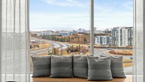 A room with a large window overlooking a cityscape, a wooden floor, and a seating area with grey cushions.