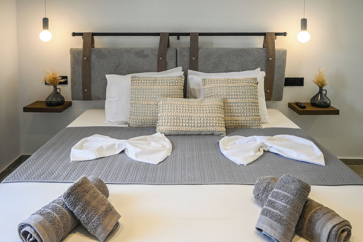 A neatly made bed with grey bedding, two pillows, and a bedside table with a vase and remote control.