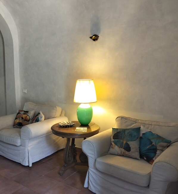 Une pièce avec deux canapés blancs, une petite table ronde sur laquelle se trouve une lampe verte, et un coussin décoratif posé sur l’un des canapés.