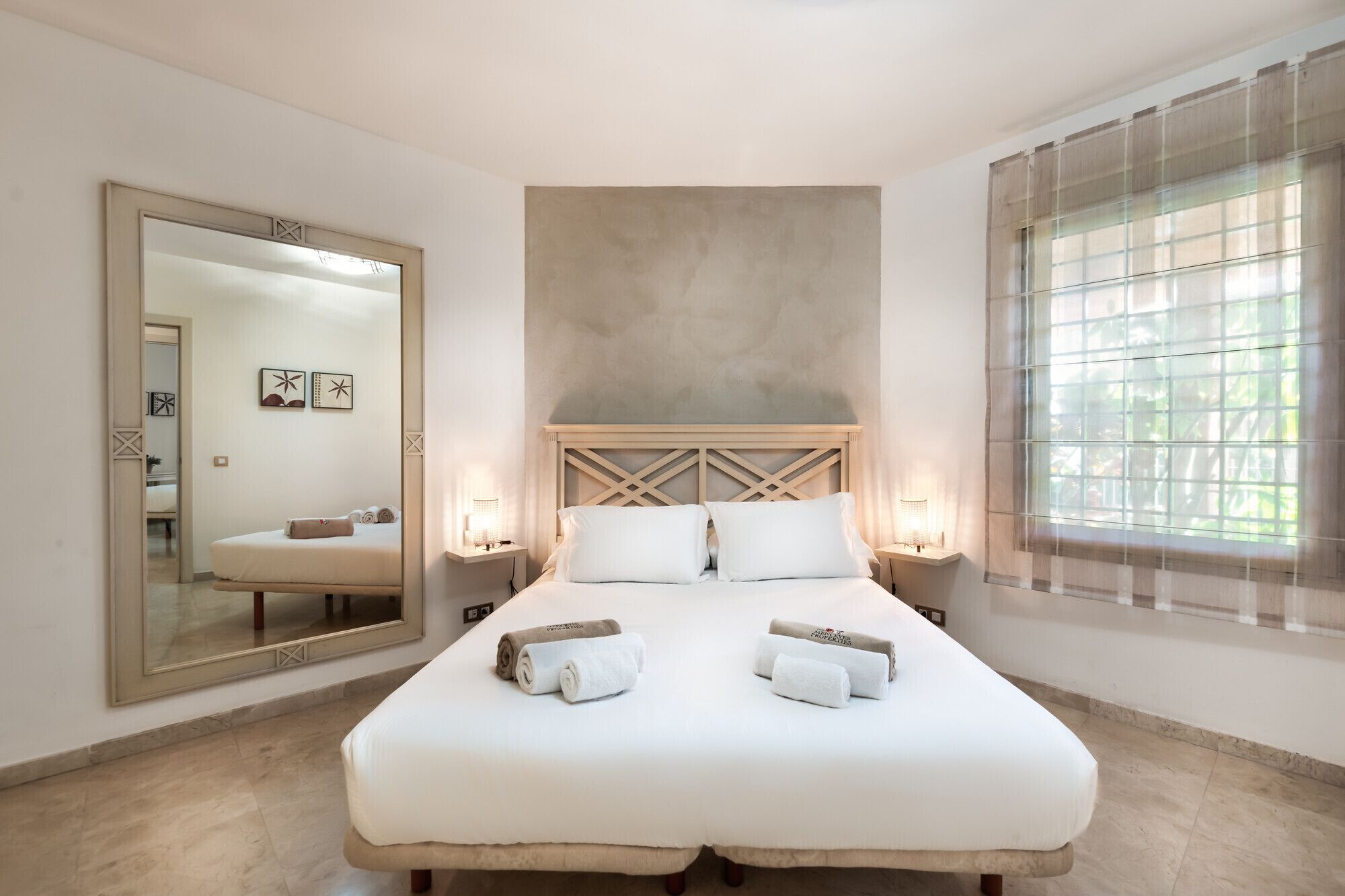 Une chambre à coucher avec un grand lit, deux tables de chevet, un miroir et une fenêtre ornée de rideaux légers.