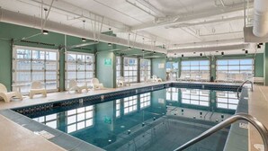Una amplia piscina interior con ventanas transparentes, suelo de baldosas y varias sillas de descanso.