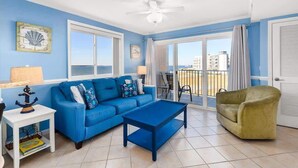 Habitación con sofá azul, sillón verde, mesita auxiliar blanca y mesa de centro azul. Hay un ventilador de techo, una ventana con vista al mar y una puerta con vista al mar.