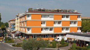 A multi-story hotel with orange and white exterior walls, a central courtyard with a tree, and a street view with pedestrians and vehicles. - Foyer in Bibione With 1 Bedrooms and 1 Bathrooms (San Michele al Tagliamento)