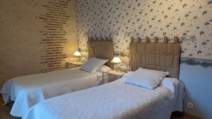 Una habitació amb dos llits individuals, cadascun amb un capçal i una taula de nit amb una làmpada. Les parets tenen paper pintat floral, i hi ha una paret de maó a un costat.
