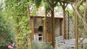 Jardí amb una pèrgola, cadires blanques i una estructura de pedra amb teulada.