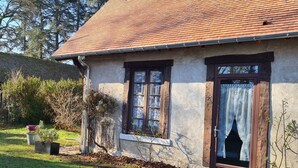 Una casa rústica con techo de tejas, puerta de madera con cortinas de encaje y un pequeño jardín con césped y plantas en macetas.