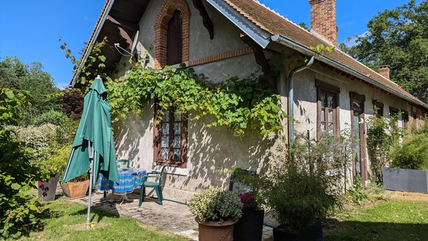 Una casa tradicional con techo inclinado, chimenea de ladrillo y un jardín con plantas en macetas y una mesa con sillas.