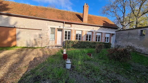 Un bâtiment en pierre de deux étages, avec un toit recouvert de tuiles rouges, une cheminée et un petit jardin agrémenté d’un seau et d’un objet blanc.