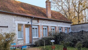 Un bâtiment en pierre avec un toit en tuiles, une cheminée en briques et un petit jardin agrémenté d’une table et de chaises.