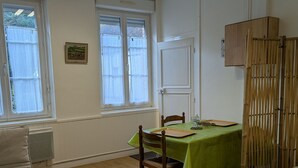 Une pièce comprenant une table à manger, des chaises, un radiateur, une fenêtre avec des rideaux, une porte et une cloison en bois.