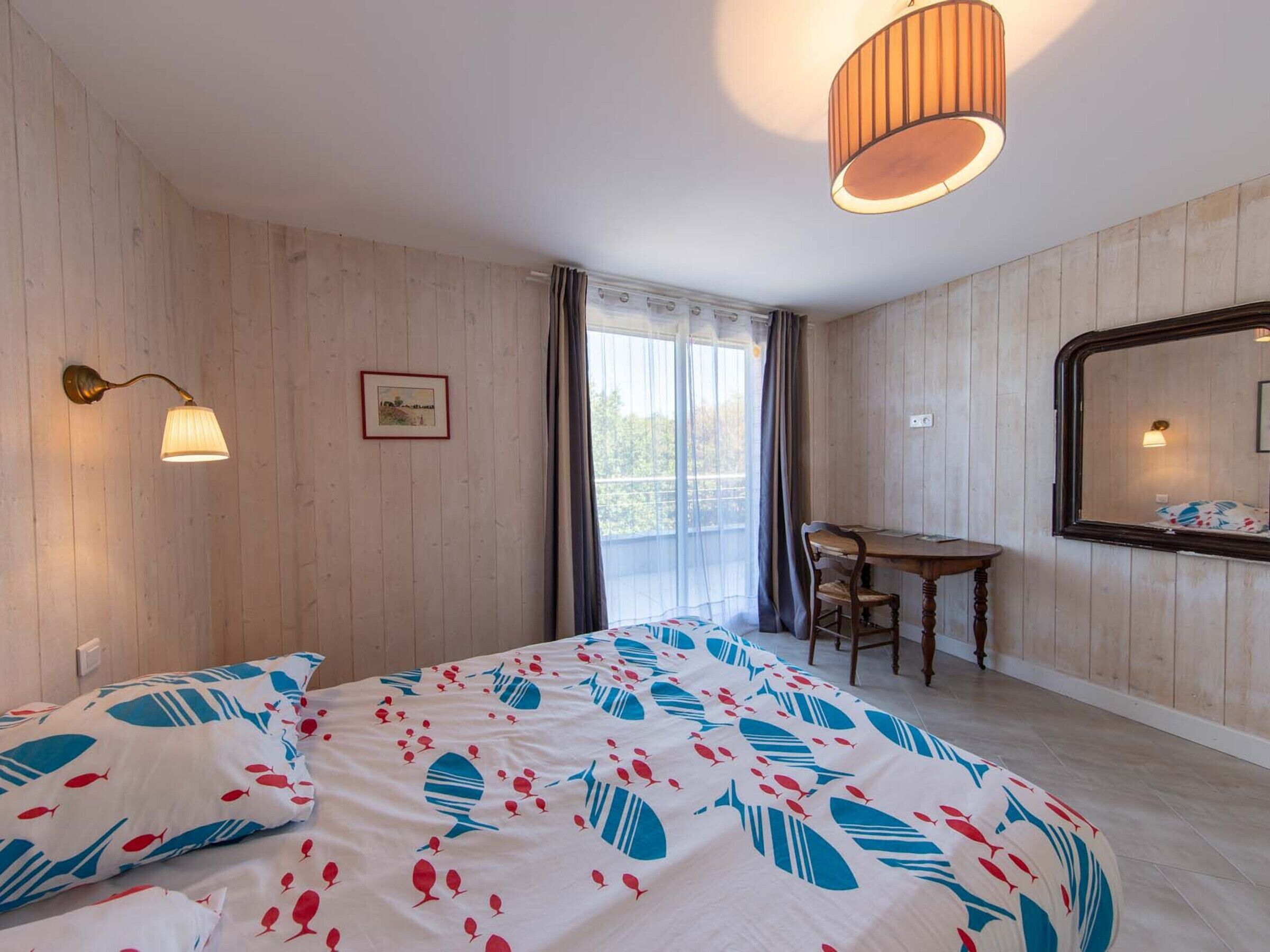 Une chambre d’hôtel comprenant un lit, un bureau, une chaise, une fenêtre avec des rideaux et une lampe fixée au mur.