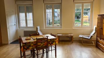 Un comedor con una mesa dispuesta para cuatro personas, rodeada de sillas de madera. Hay dos ventanas con persianas, un radiador y un aparador de madera al lado.