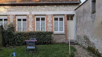 Un edificio de piedra con fachada de ladrillo, una puerta y ventanas. Hay un pequeño espacio con césped, una silla de metal y una mesa.