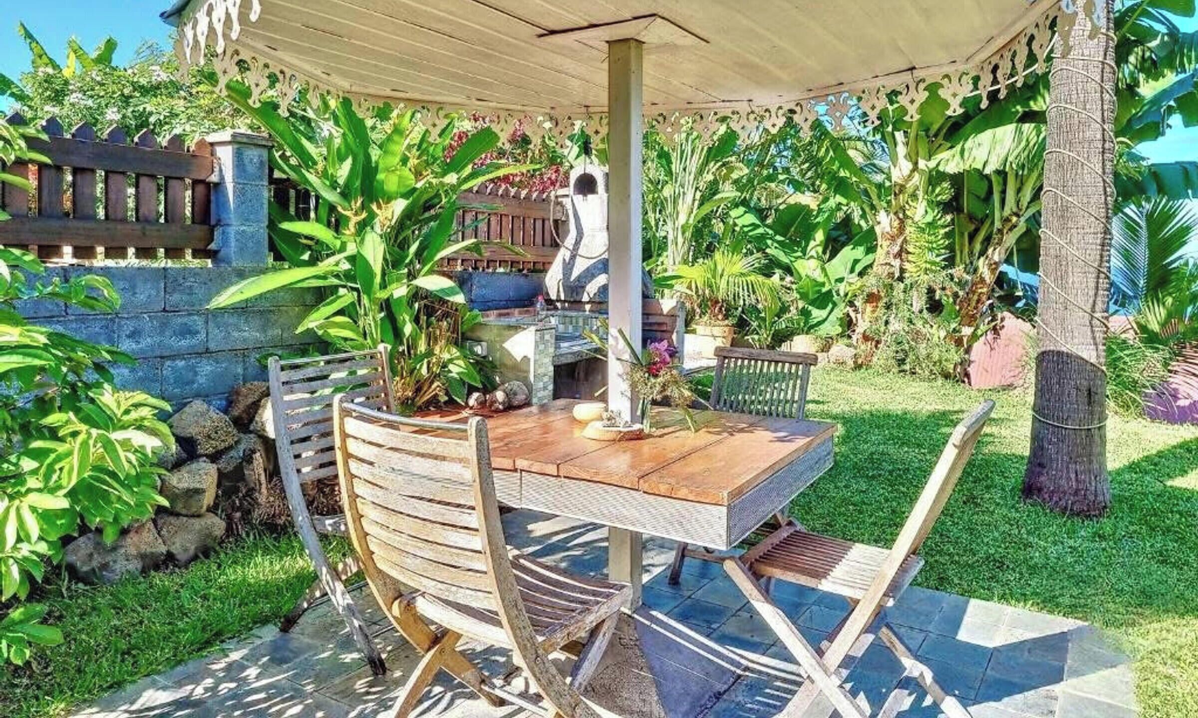 An outdoor patio with a wooden table and chairs, surrounded by lush greenery and a decorative fence.