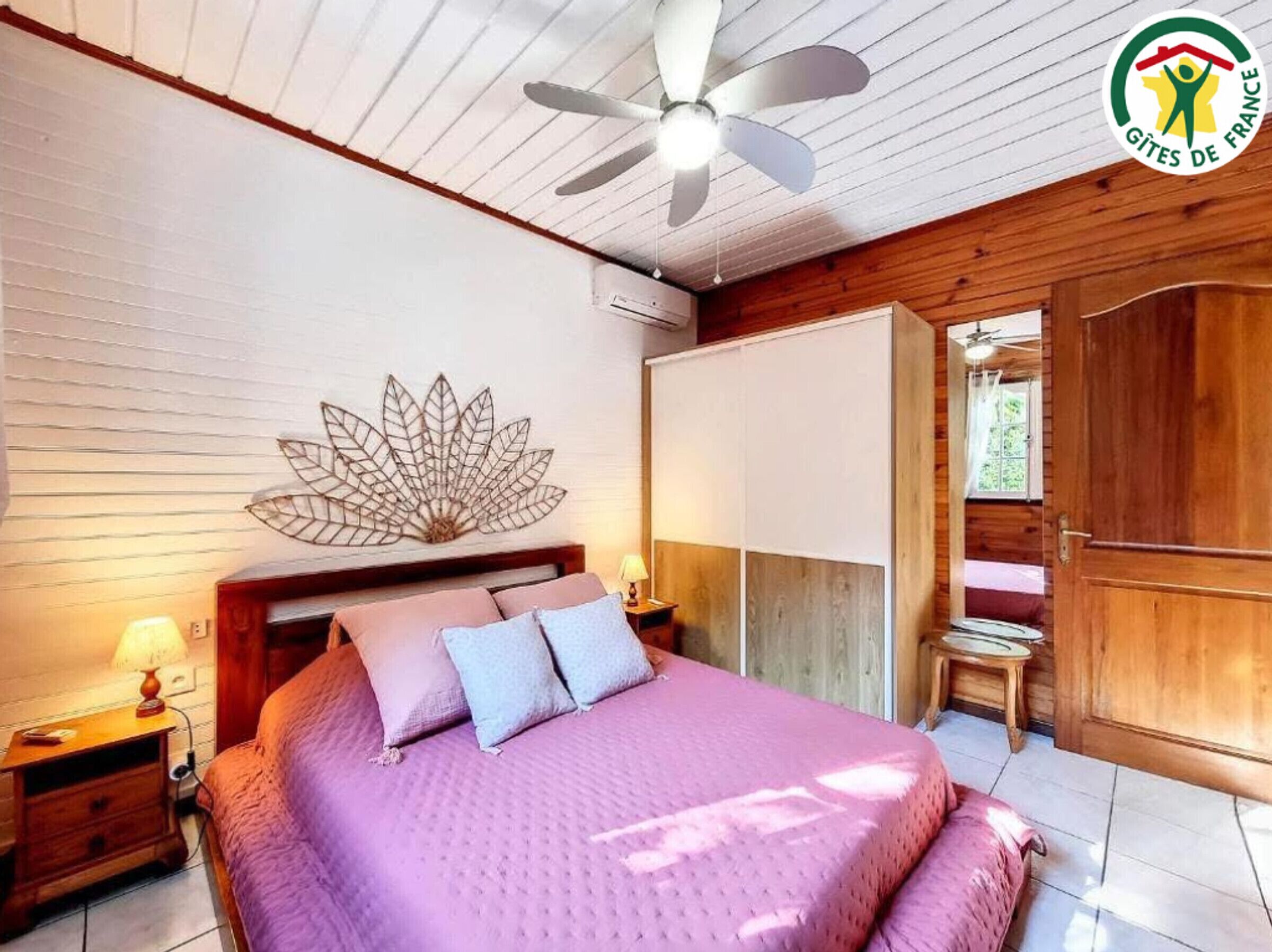 A bedroom with a bed, a ceiling fan, a wall-mounted decorative leaf, a wooden wardrobe, and a door leading to another room.