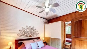 Een slaapkamer met een bed, een plafondventilator, een decoratief blad aan de muur, een houten garderobe en een deur die naar een andere kamer leidt.