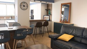 Una sala de estar moderna con un sofá de cuero negro, una mesa de comedor de madera con sillas y una cocina visible a través de una puerta abierta.
