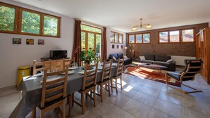 A spacious living area with a dining table, chairs, a sofa, and a TV. Large windows provide natural light and views of greenery outside.