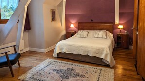 A bedroom with a sloped ceiling, a bed with two pillows, two bedside tables with lamps, a wooden chair, and a patterned rug.