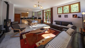 A modern living room with a sofa, a coffee table, and a fireplace. There is a kitchen area with wooden cabinets and a dining table. Large windows provide natural light and views of greenery outside.