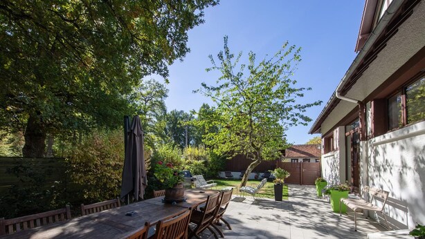 An outdoor patio with a wooden table and chairs, surrounded by greenery and a house in the background.