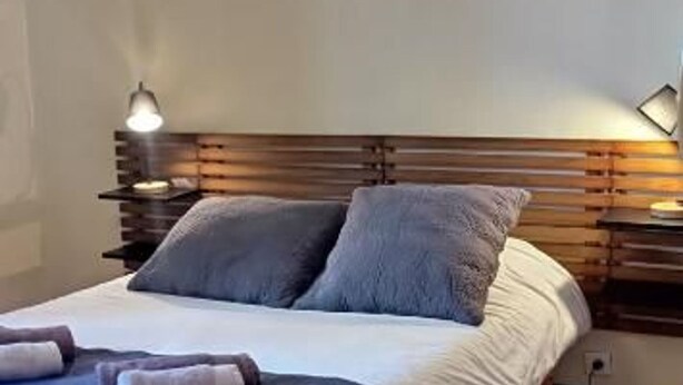 Un lit bien fait, avec des oreillers et une couverture, une tête de lit en bois, des tables de chevet avec des lampes et un mur sobre.