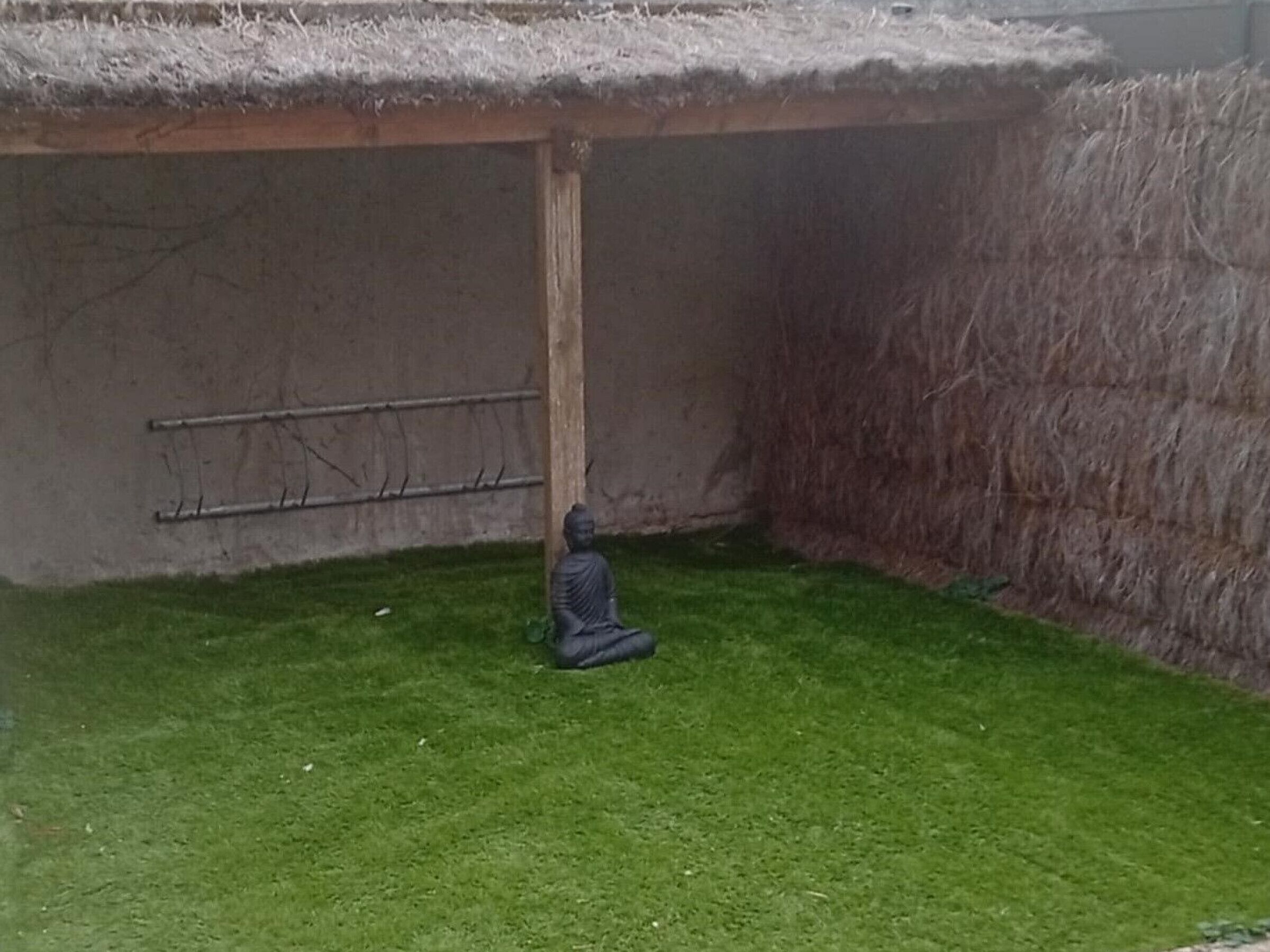 A statue of a seated figure in a grassy area with a thatched roof structure and a metal rack against a wall.