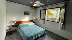 Une chambre à coucher avec un lit, un ventilateur de plafond, une fenêtre donnant sur des plantes et une petite table de chevet avec une lampe.