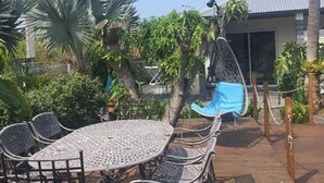Une terrasse extérieure avec une table et des chaises en métal, entourée d’une végétation luxuriante et agrémentée d’un fauteuil suspendu.