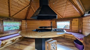 A wooden cabin interior with a central table, benches, and a black stove.