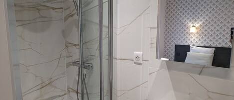 Un bagno moderno con doccia vetrata, un lavandino bianco con dispenser di sapone e uno specchio che riflette un letto con testiera decorata.