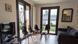 Ein Wohnzimmer mit Fernseher, schwarzen Sesseln, einer Couch und einem Fenster mit Blick auf ein Haus.