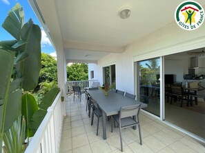 A balcony with a dining table and chairs, a sliding glass door leading to a kitchen, and a view of lush greenery.