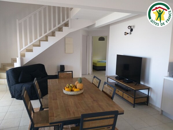 A modern living room with a wooden dining table, a black sofa, a television, and a staircase leading to another room.
