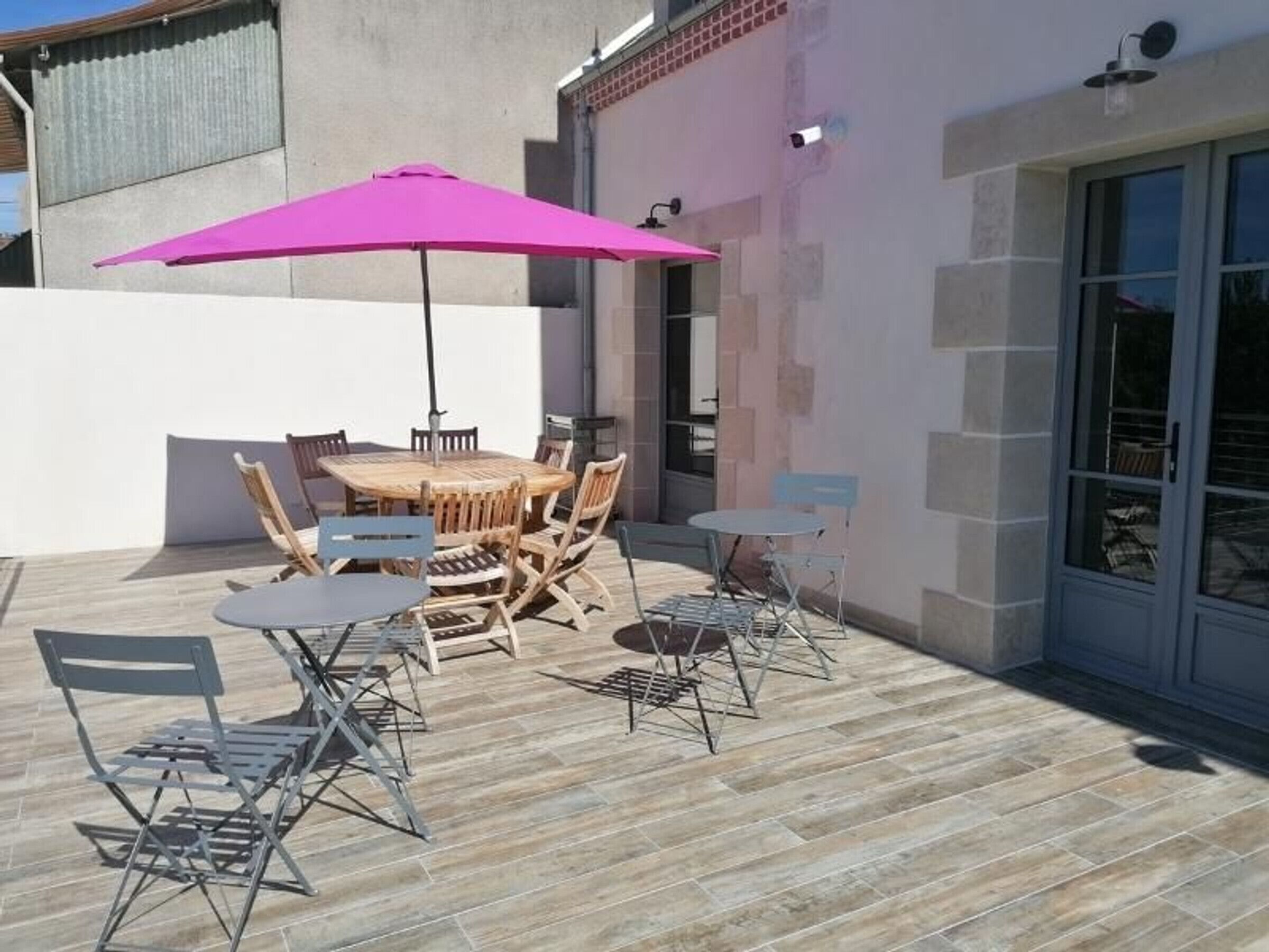 Une terrasse avec une table ronde en bois et des chaises, un parasol rose et une porte vitrée donnant sur un espace abrité.