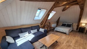 A cozy loft-style room with a dark blue sectional sofa, a wooden coffee table, a small round rug, a bed with patterned bedding, and two skylights allowing natural light. - Large Cottage in Briare - Pets and Private Parking (Briare)