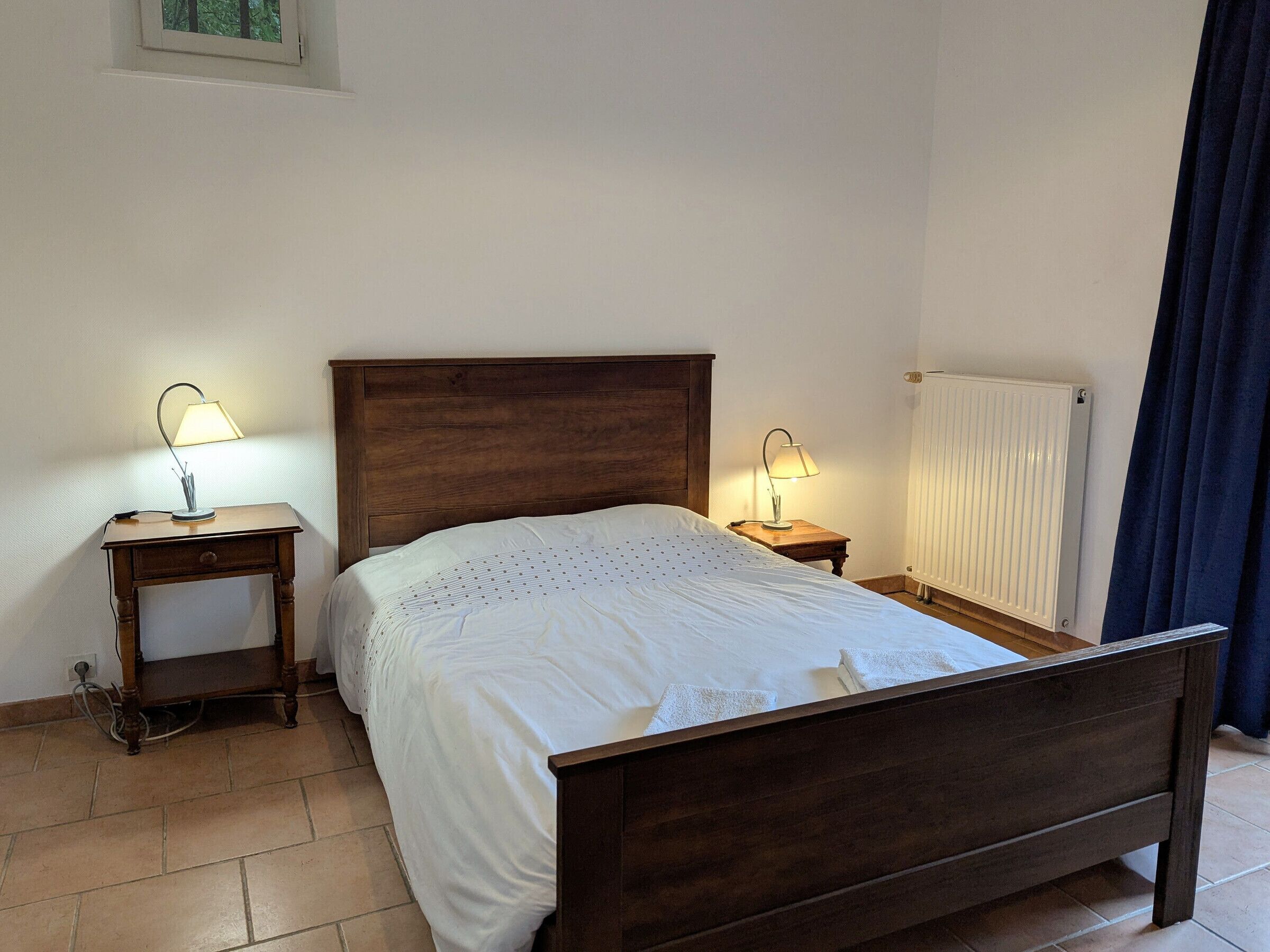 Une chambre à coucher avec un lit en bois, deux tables de chevet avec des lampes, un radiateur et une fenêtre donnant sur des arbres.